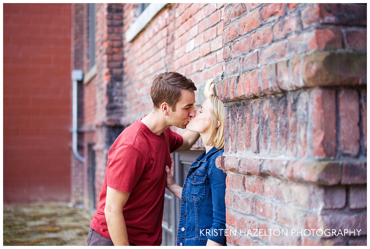 Engaged couple kissing next to brick building