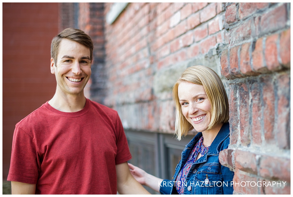 Engaged couple smiling at the camera next to a brick industrial building
