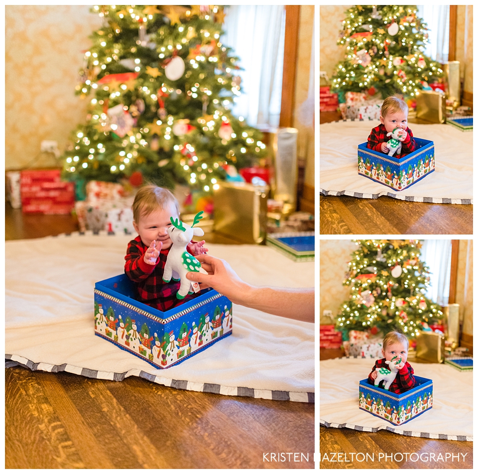 Baby in a box in front of a Christmas tree chewing on a plush reindeer