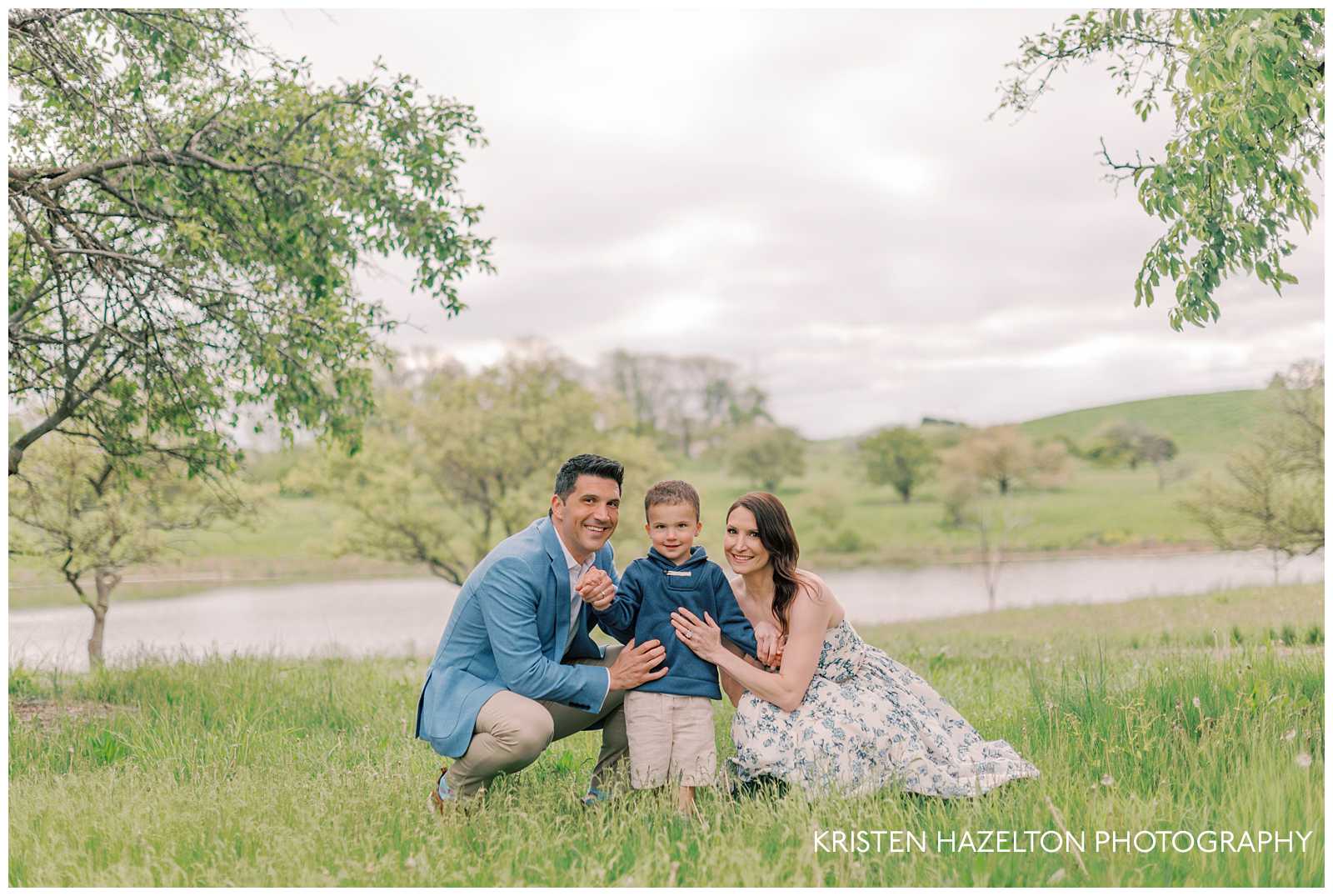 Family of three on a grassy hill overlooking a pond in the spring for their Morton Arboretum photos