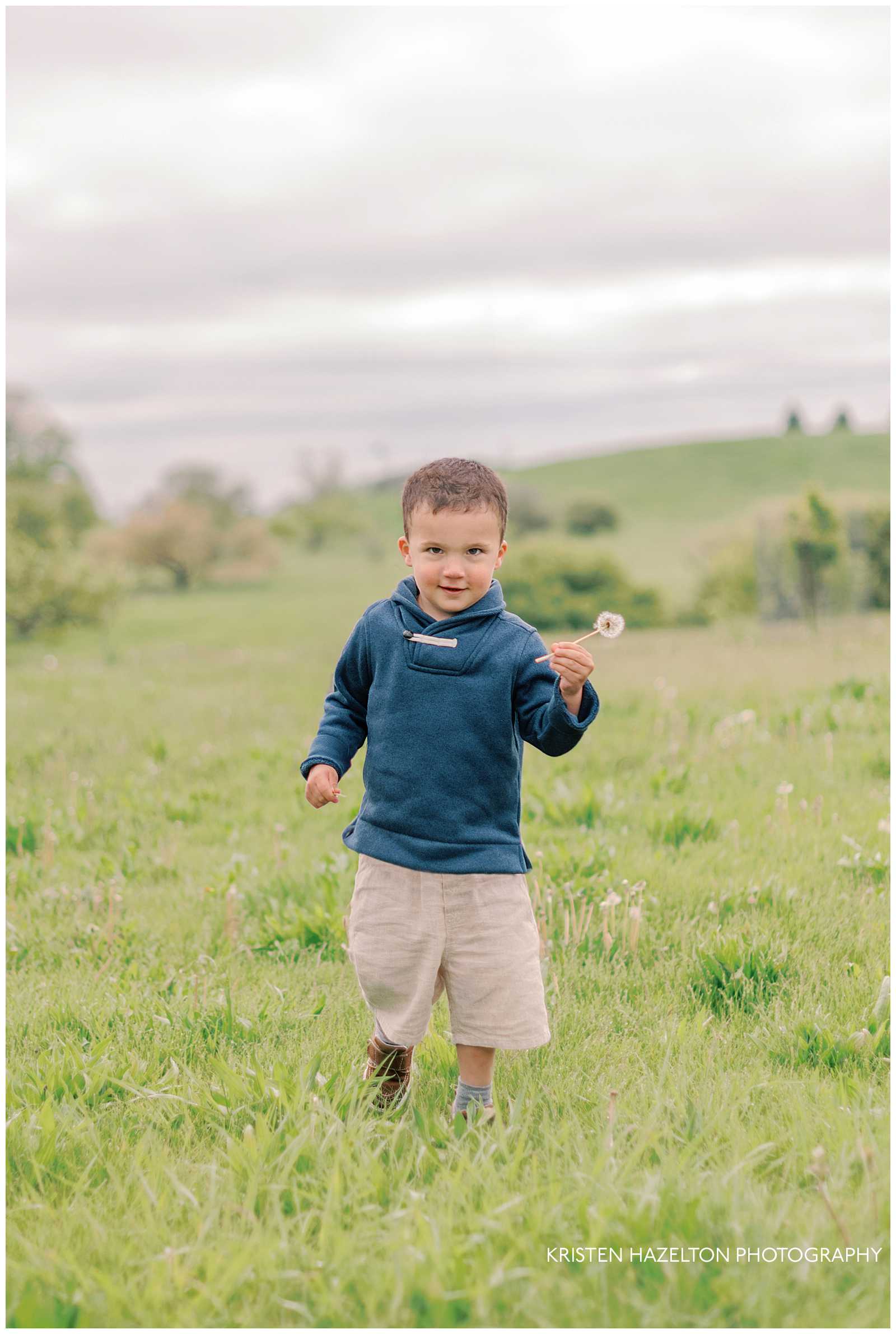 Little boy in navy sweatshirt and white linen pants running across a field while holding a dandelion