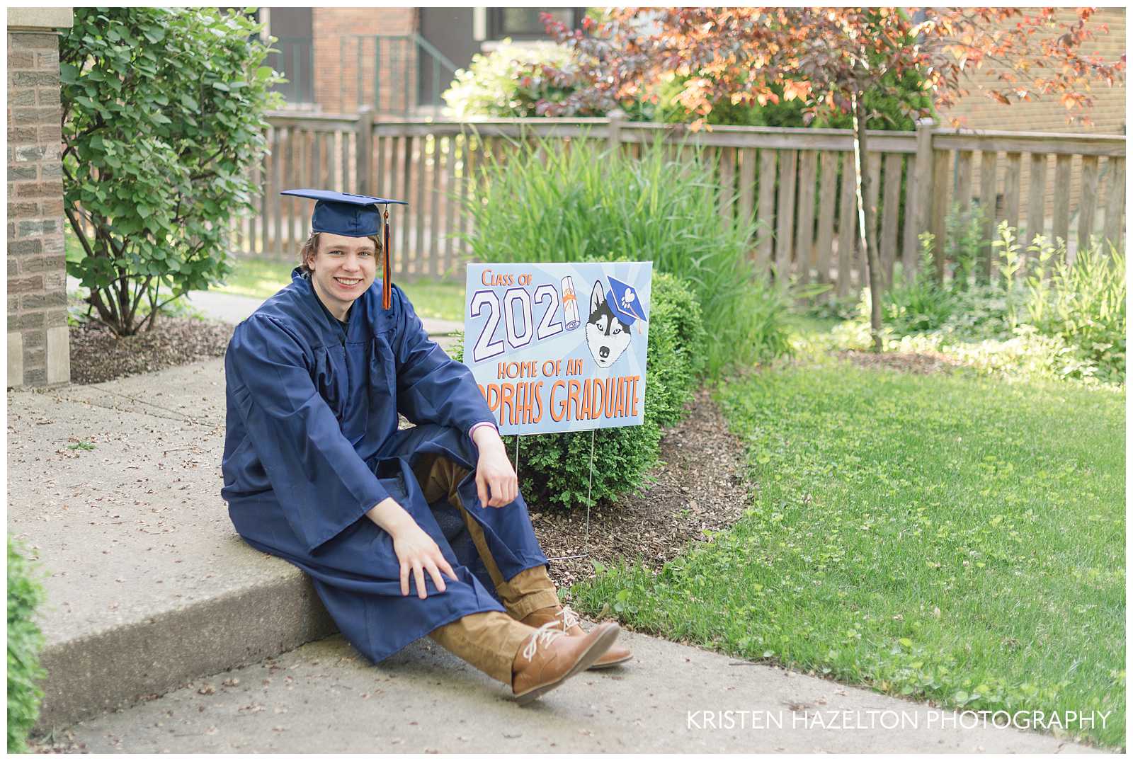 High school senior boy wearing cap and gown, sitting on a step next to a sign that reads "Class of 2021 Home of an OPRFHS Graduate."