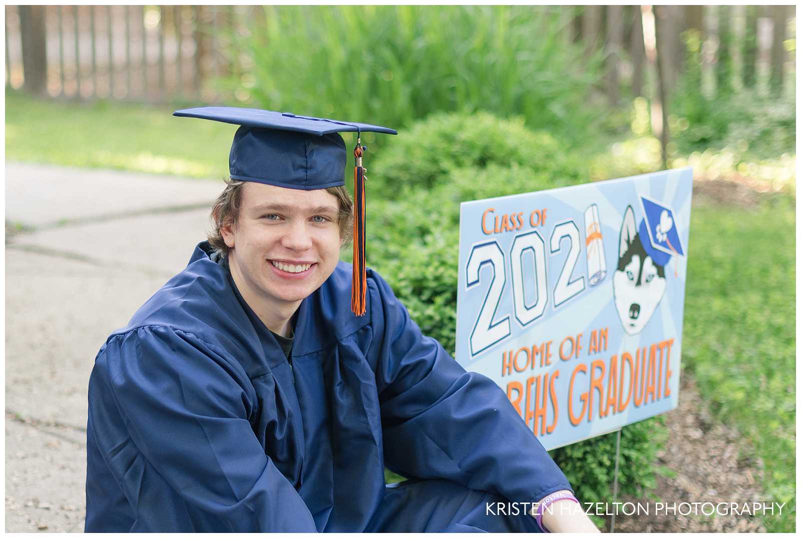 High school graduate sitting next to a "Class of 2021 Home of an OPRFHS Graduate" sign at an Oak Park, IL cap and gown photo shoot