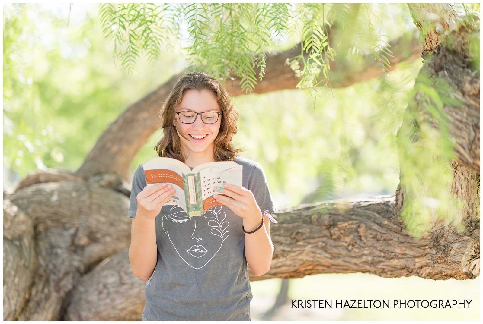 Girl reading a book and in front of a tree and smiling for her Livermore Library Senior Portraits