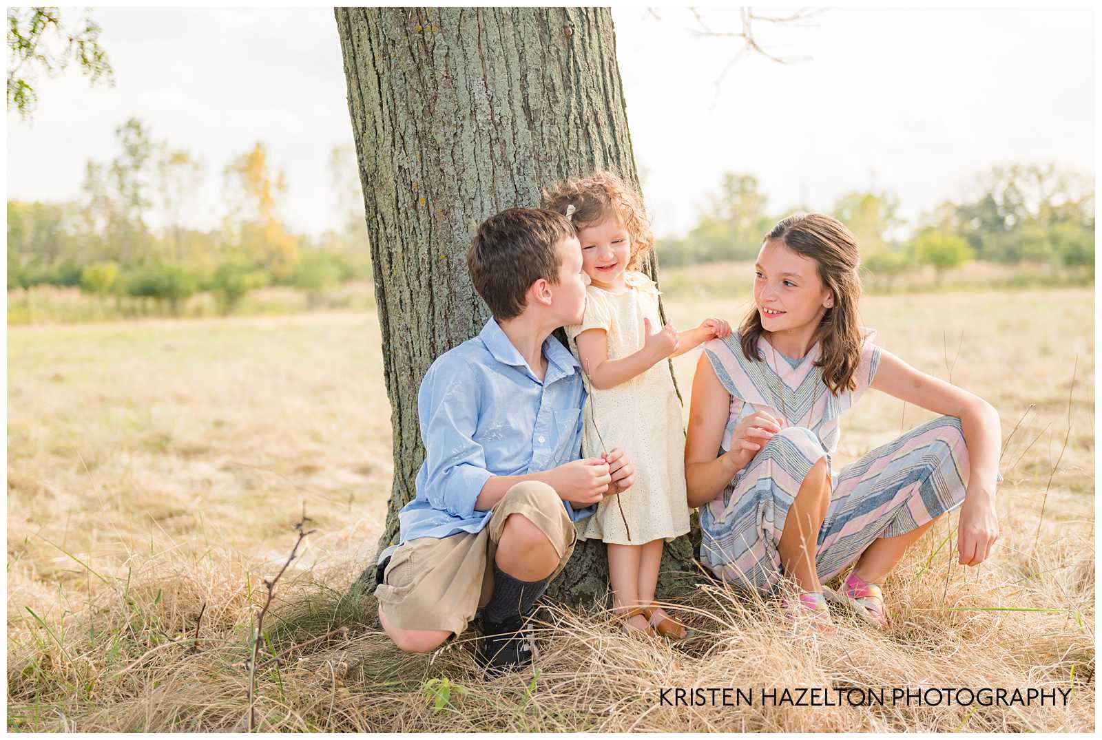 Three young siblings leaning against a tree