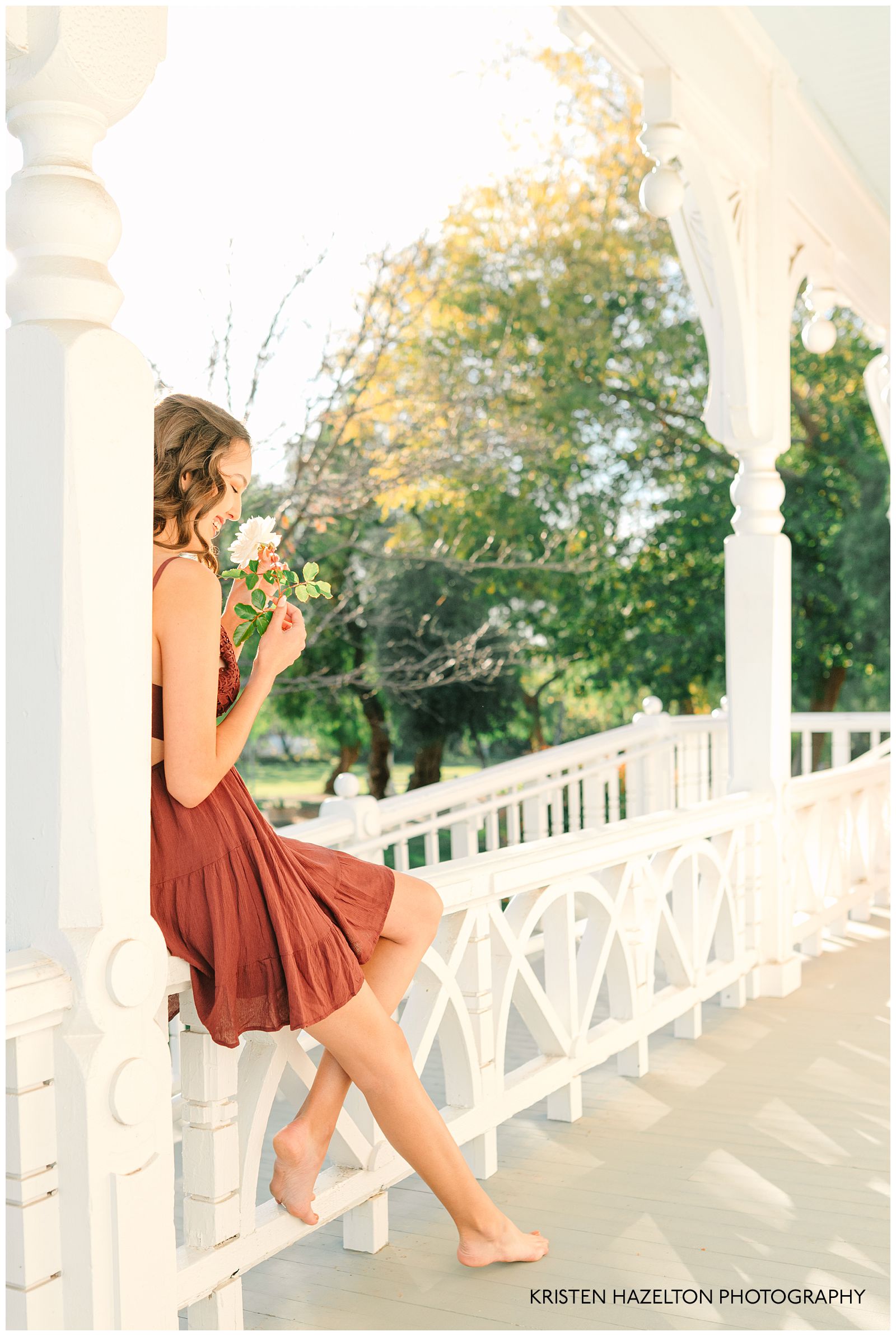 Girl in a red halter dress smelling a rose on the front porch of the Ravenswood Historical Site in Livermore, CA