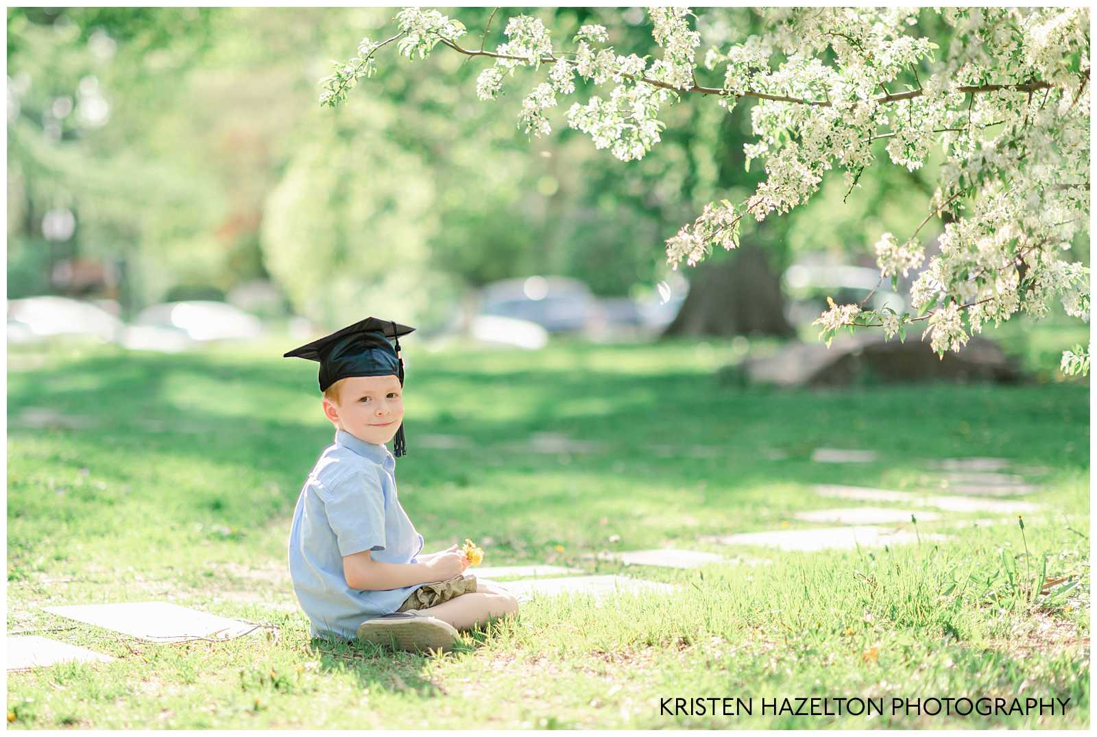 Young boy seated under a white flowering tree, wearing a graduation cap
