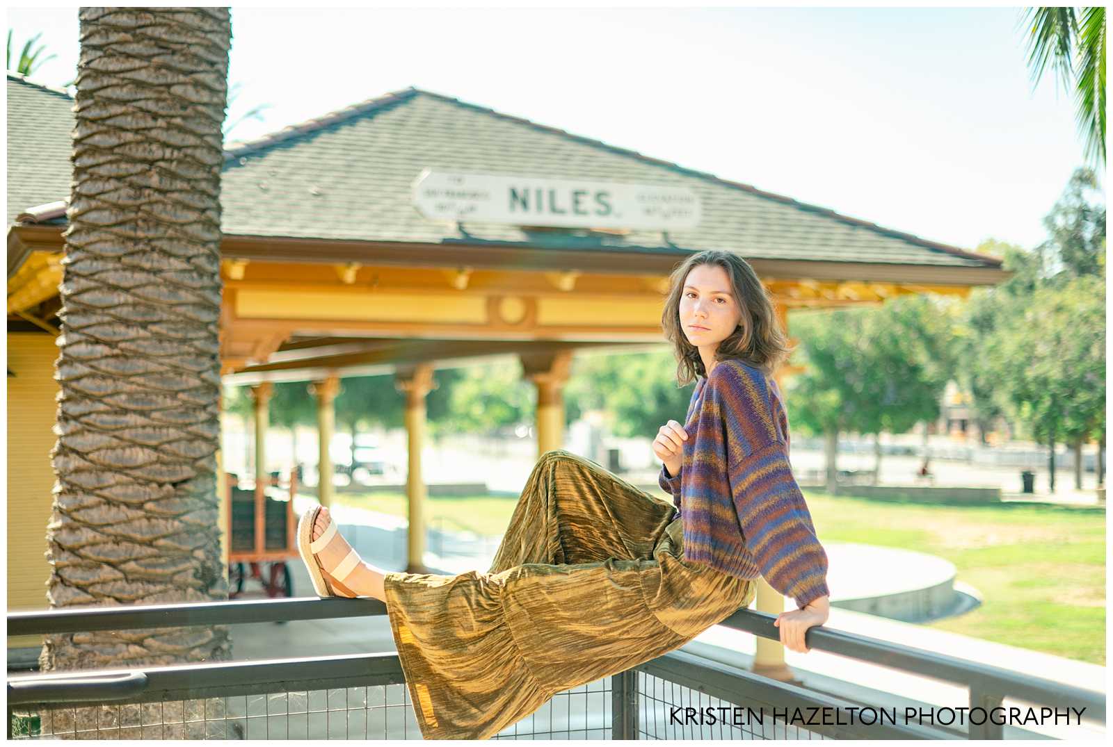 Girl wearing a striped cardigan and yellow velvet pants seated on a railing at a train station in Niles, CA