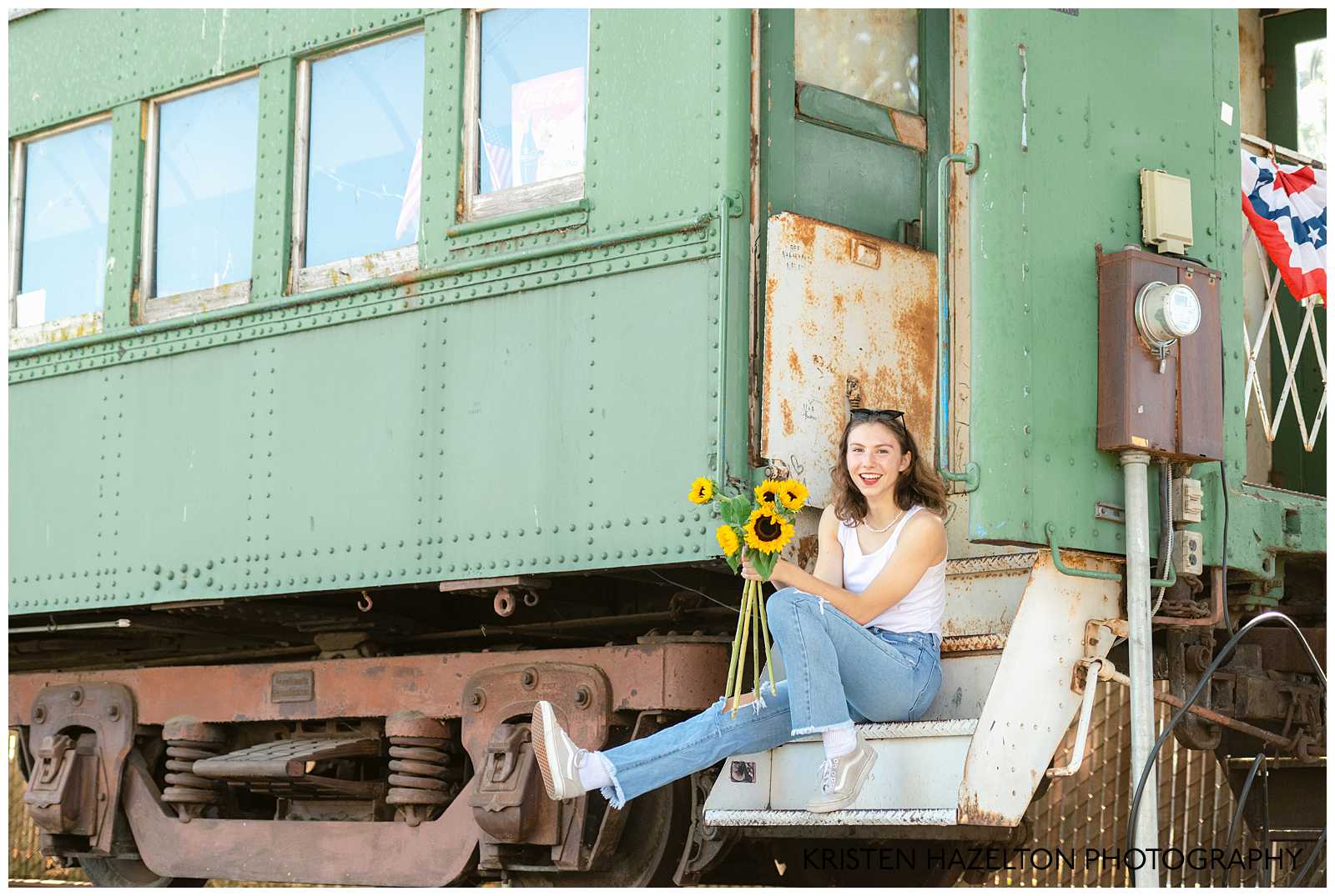 High school senior pics on a train by San Jose, CA Senior Photographer Kristen Hazelton. A girl wearing a white shirt and blue jeans sits on train  car steps while holding a bouquet of sunflowers