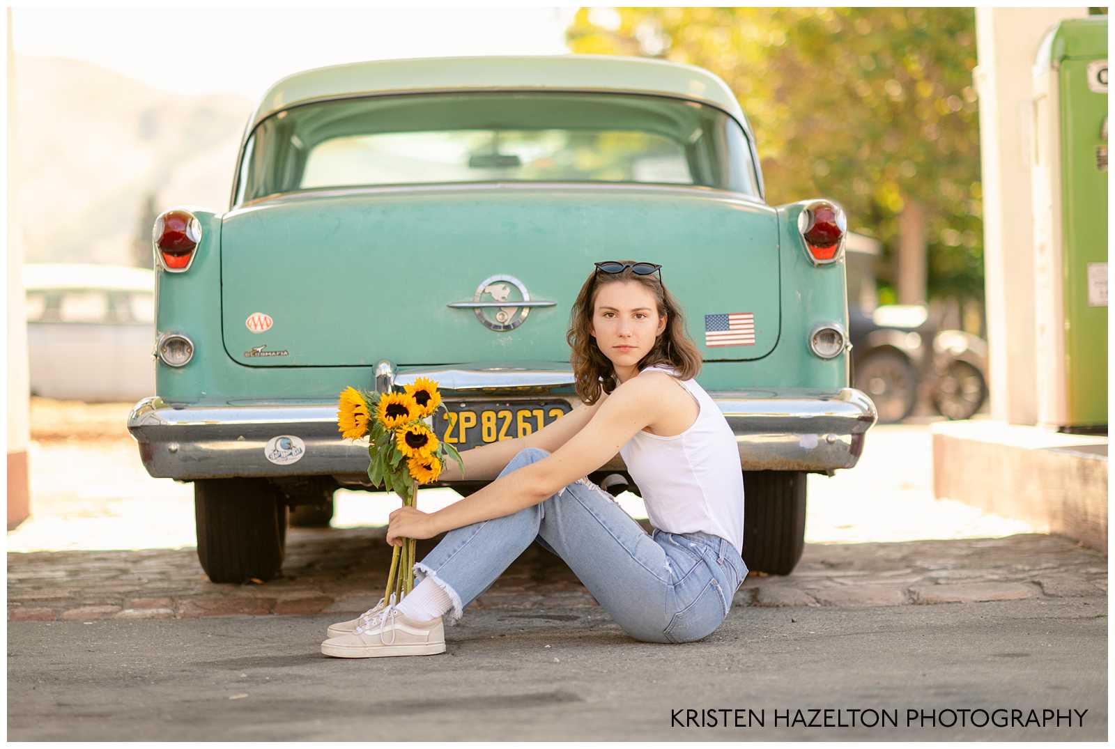 Gas station senior photos with a retro vibe: senior with sunglasses on her head, white tank top, blue jeans, and sunflowers, seated next to a teal classic car