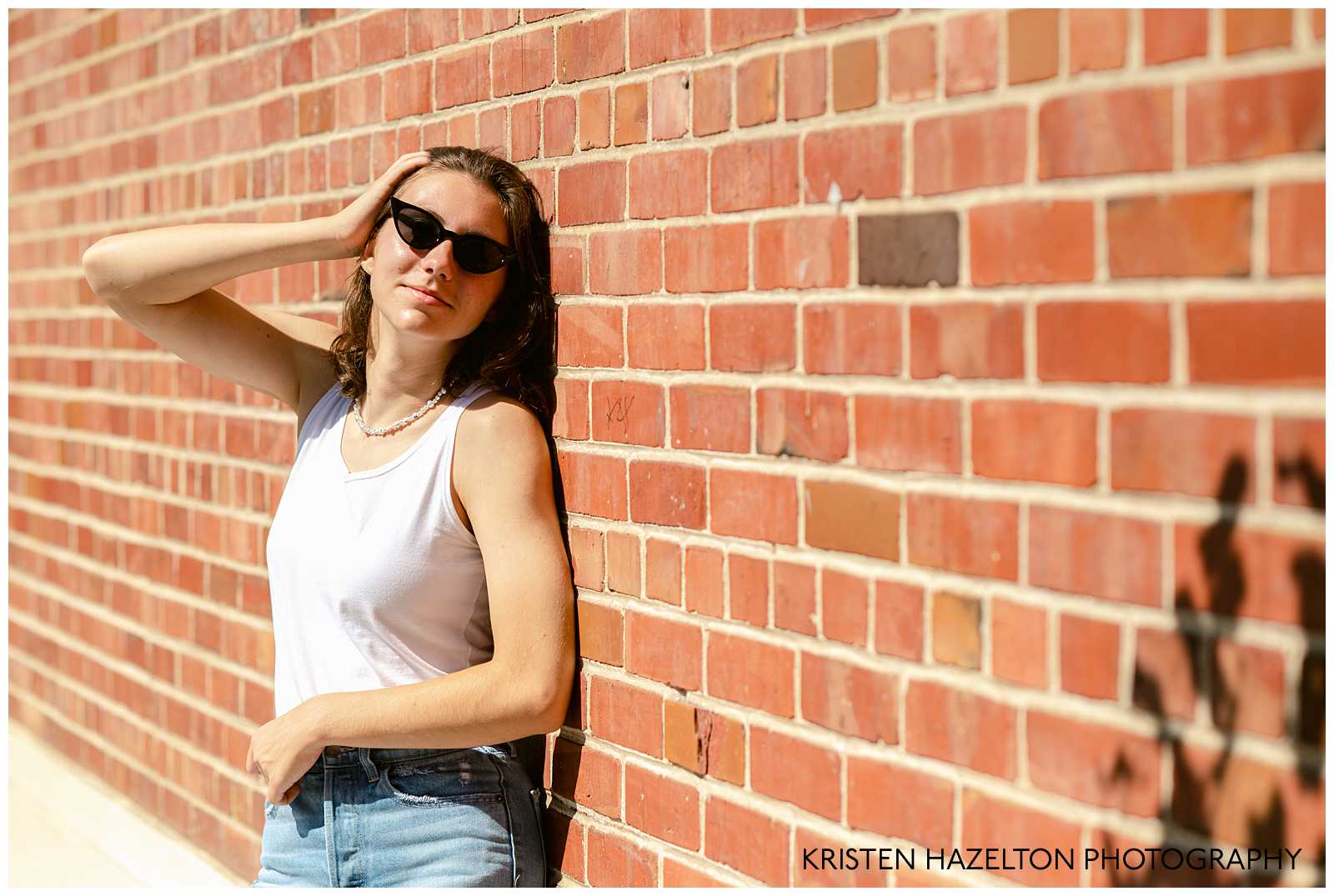 High school senior photo of a girl wearing a white tank top, blue jeans, and sunglasses, leaning against a brick wall by San Jose, CA Senior Photographer Kristen Hazelton 