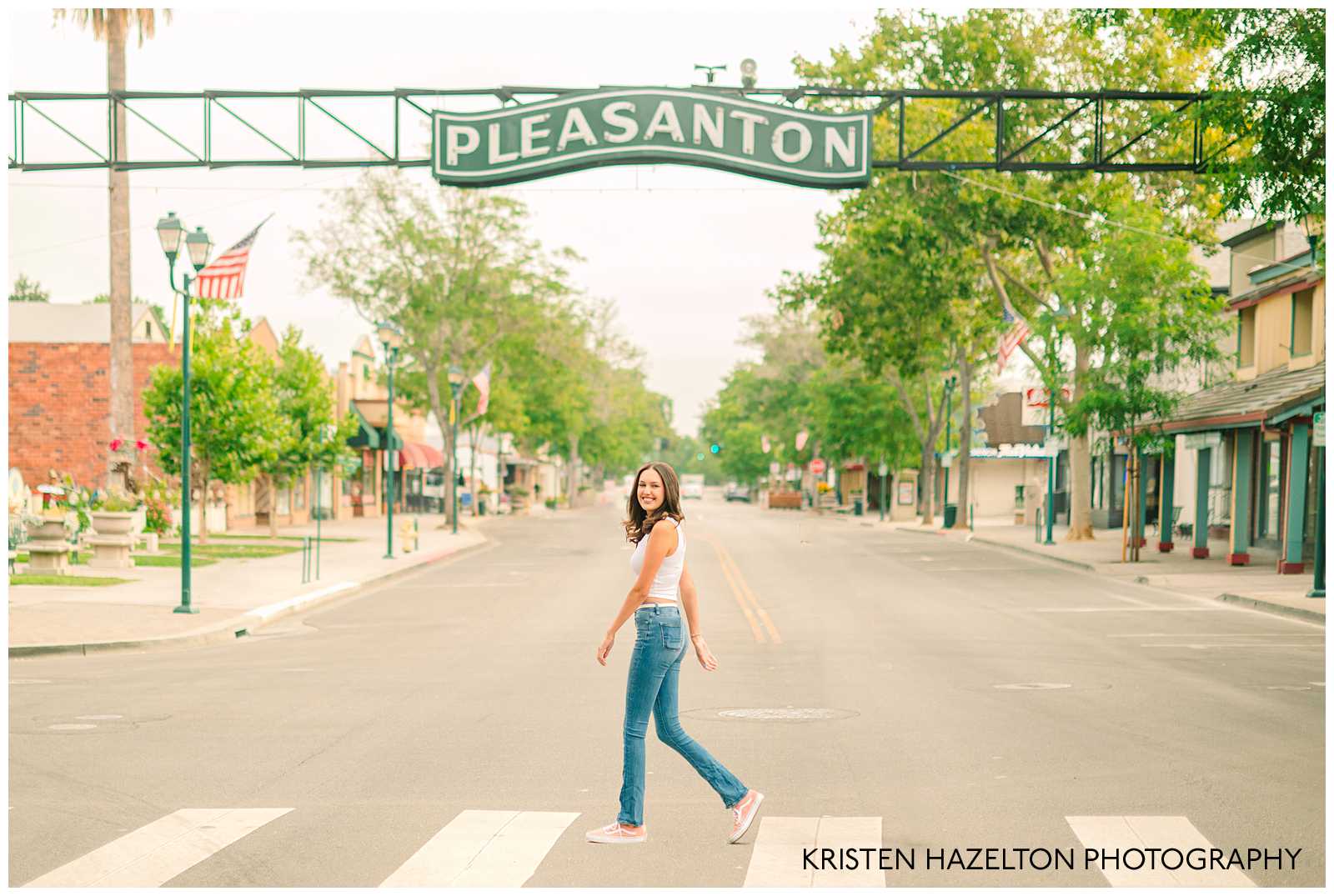 Girl wearing blue jeans and a white crop top walking across main street in Pleasanton, California by Pleasanton CA senior photographer Kristen Hazelton
