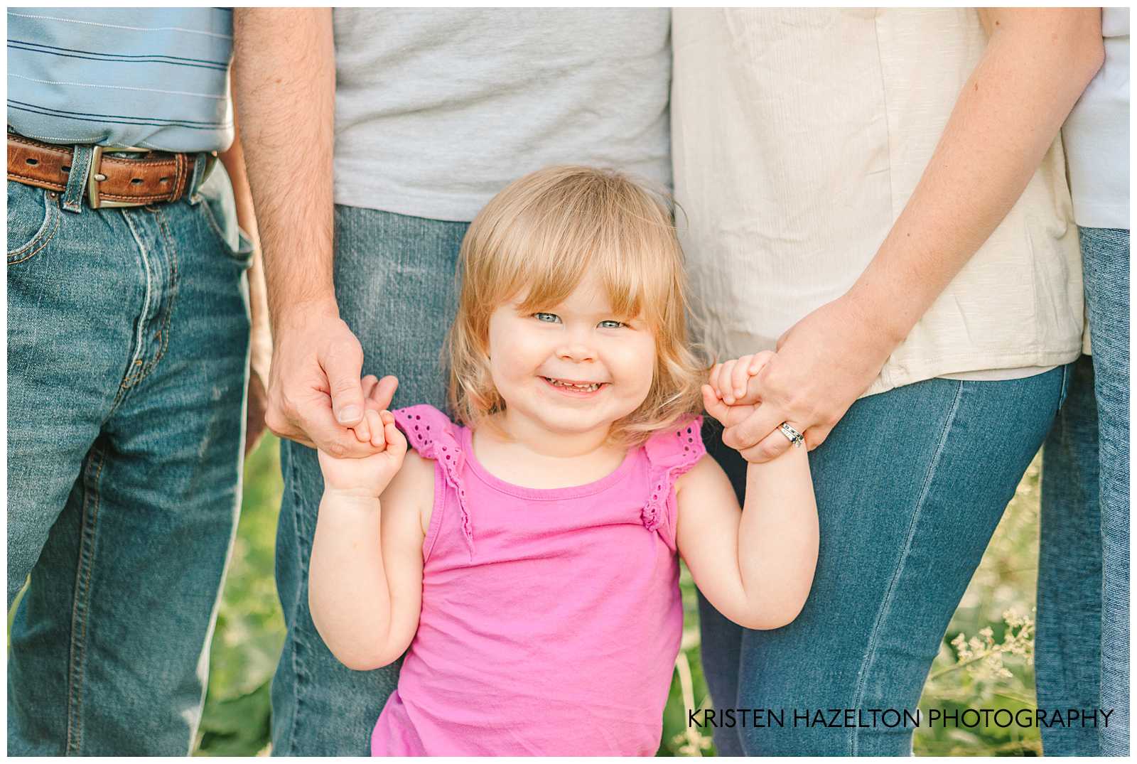 closeup of a toddler girl with a pink shirt holding her parents' hands.