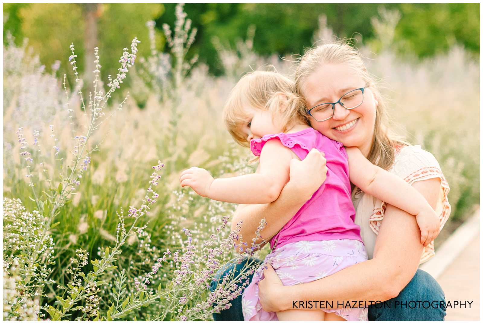 Mom hugging her toddler daughter who is touching purple Russian Sage flowers. 