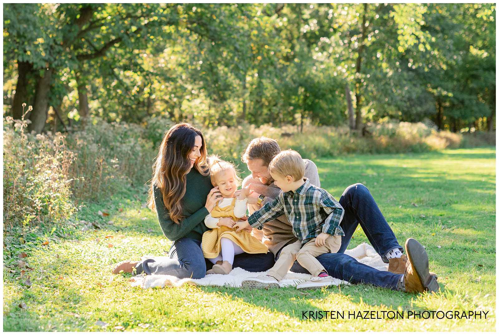 Family of four sitting on a blanket on some grass, tickling the youngest child