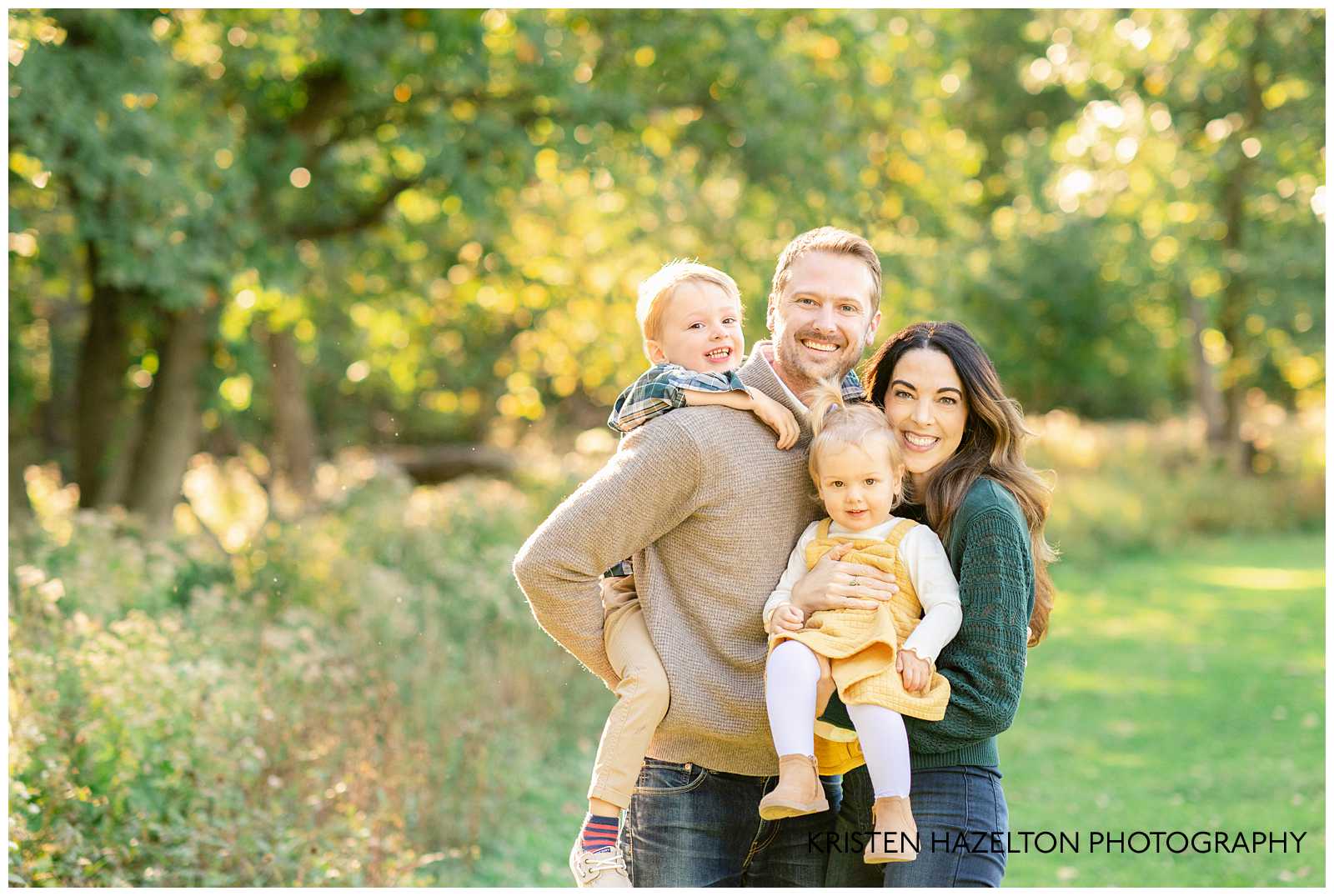 Family of four with two toddler children, smiling in a grassy field by Elmhurst family photographer Kristen Hazelton