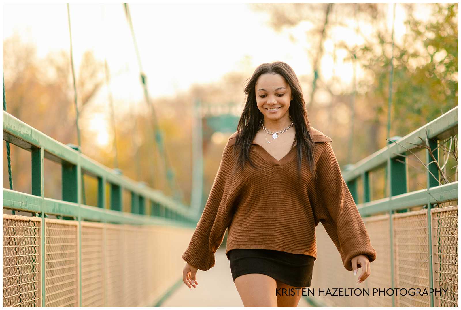 OPRF High school senior photos of a girl in a brown sweater and black mini skirt walking on a green suspension bridge.