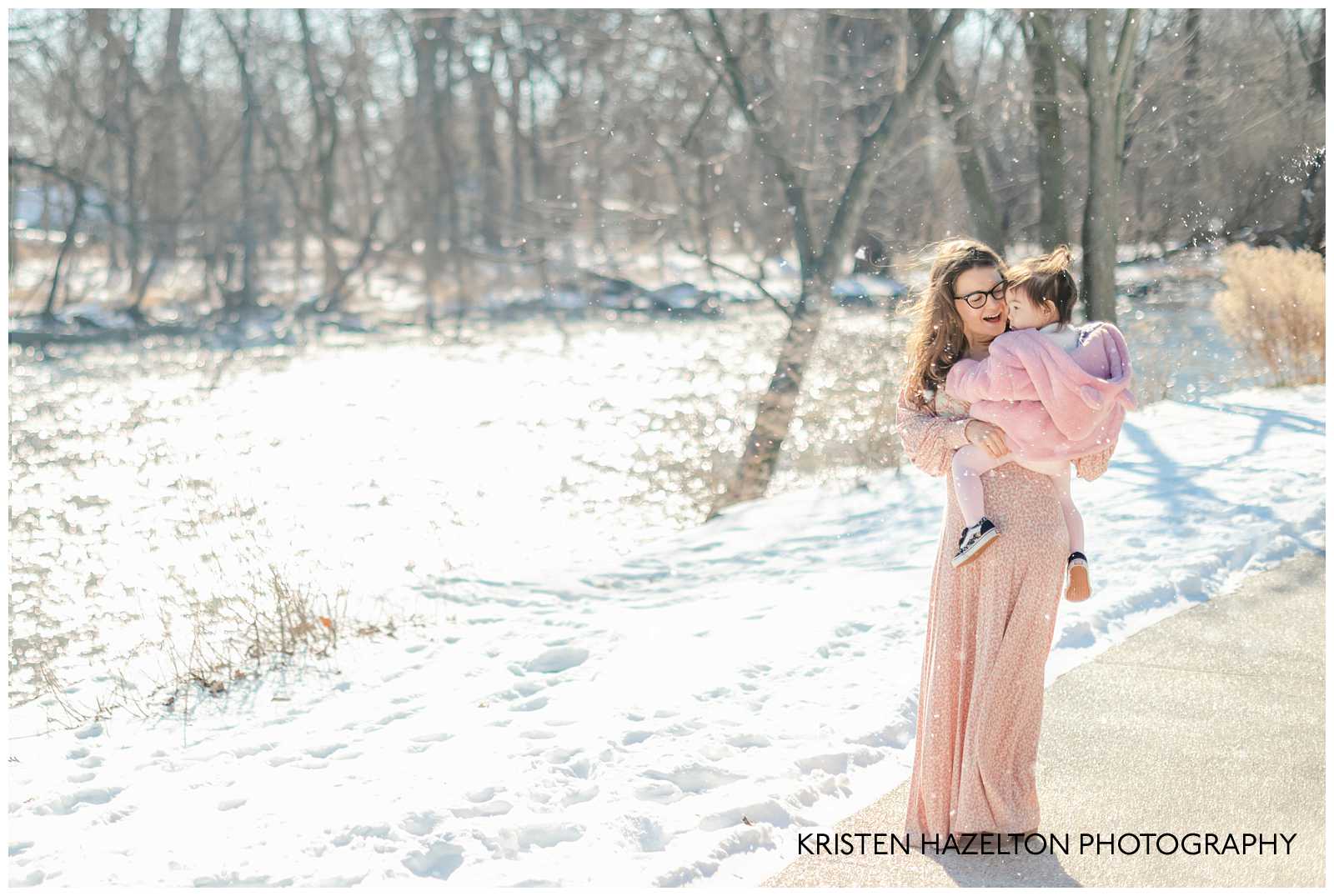 Mom wearing glasses and pink dress holding her toddler daughter on a snowy day next to a river. Winter photoshoot ideas by Kristen Hazelton