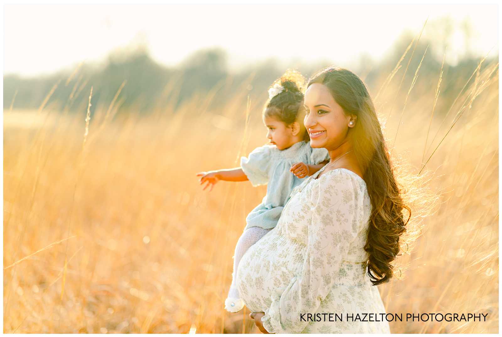 Pregnant woman wearing a light colored dress in a field of yellow grass holding her toddler daughter