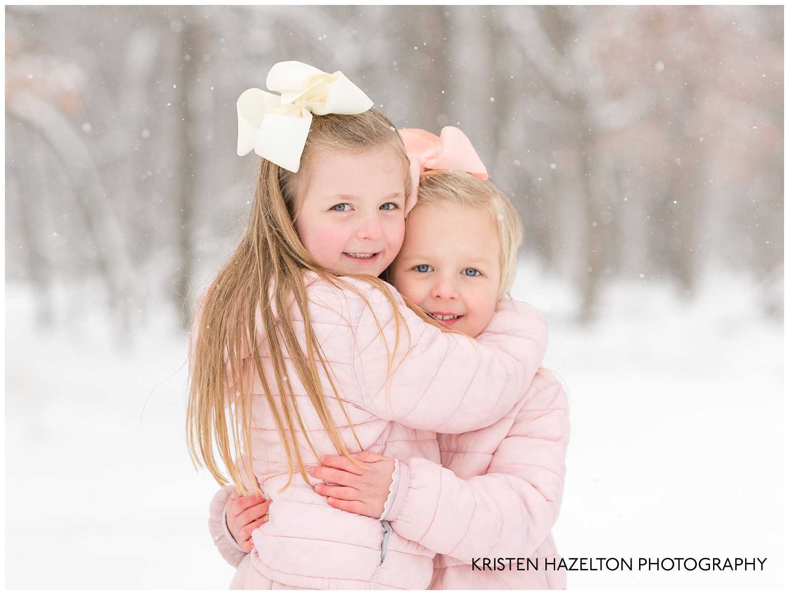 Two young sisters wearing pink jackets and hugging each other in the snow