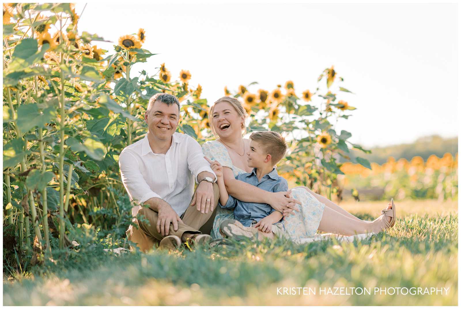 Family of three sitting in a field and laughing during their sunflower photos