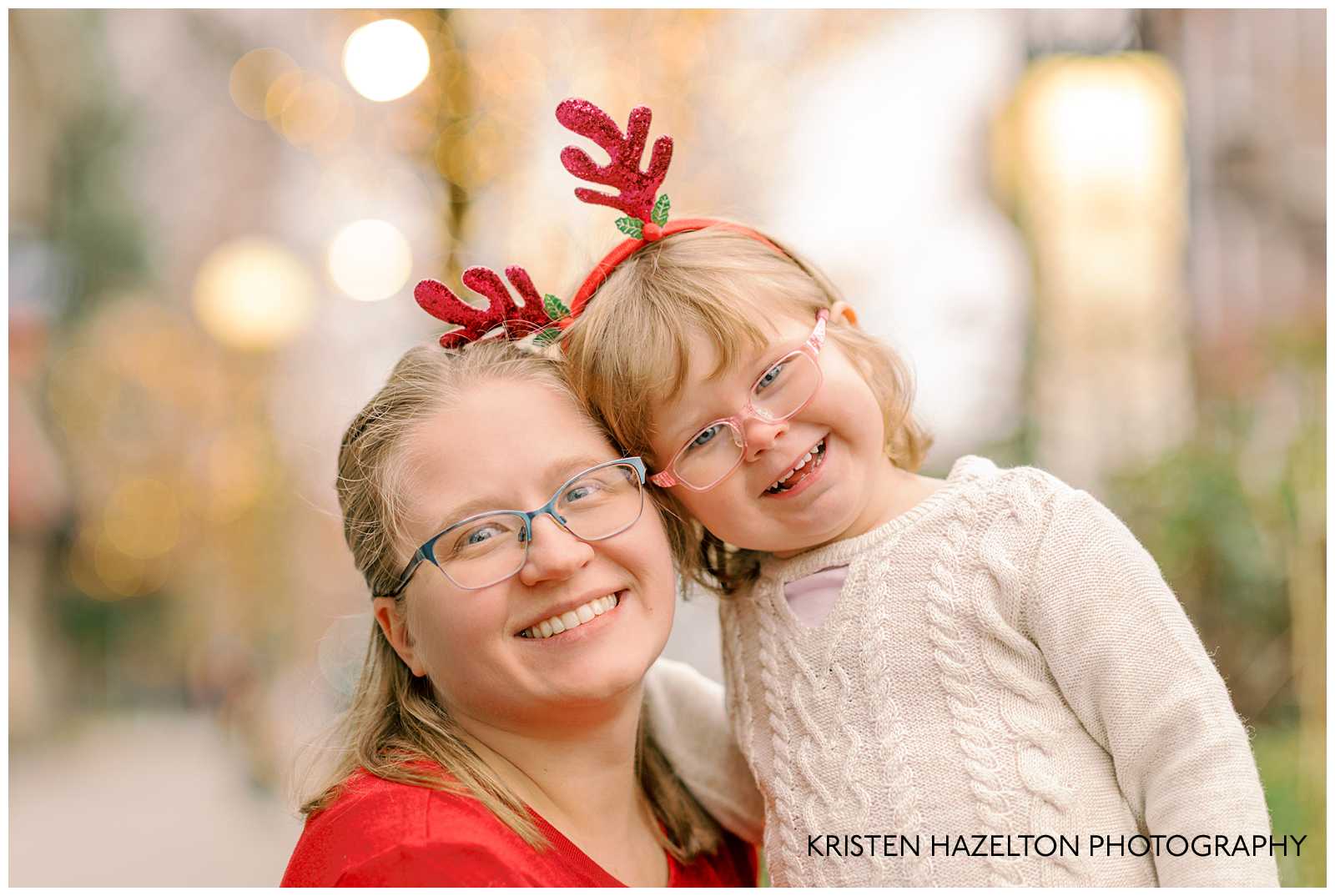 Closeup photo of a woman in a red dress with blue glasses and her toddler daughter in a white sweater dress and red reindeer antler headband.
