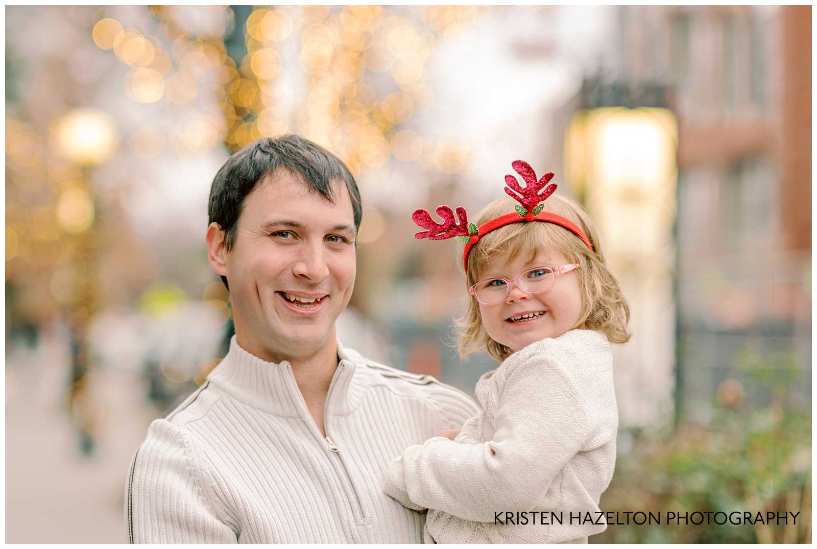 Dad in white sweater hugging his young daughter wearing red reindeer antler headband.