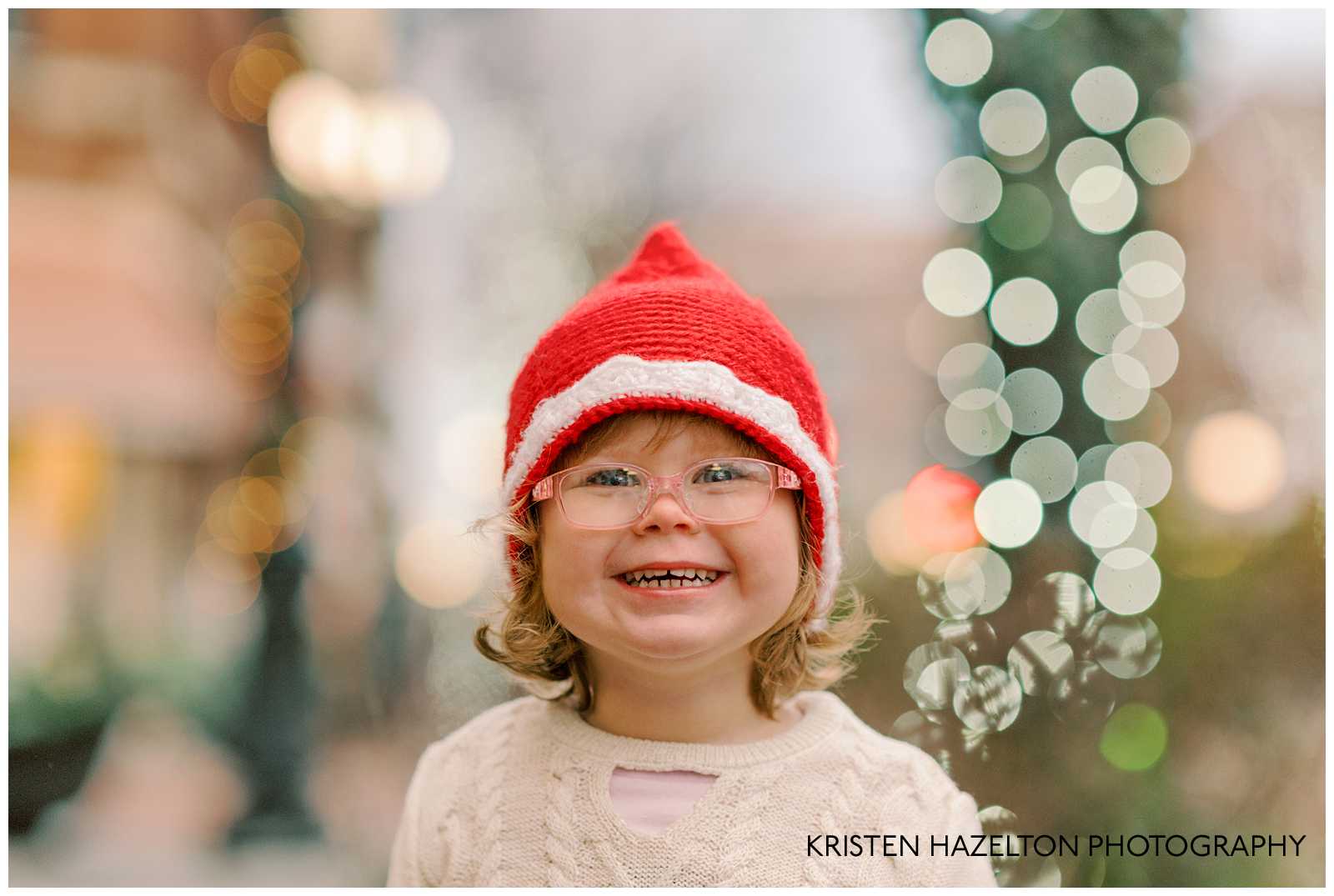 Little girl in a red Santa hat and white sweater dress grinning as she looks at holiday lights in Chicago.