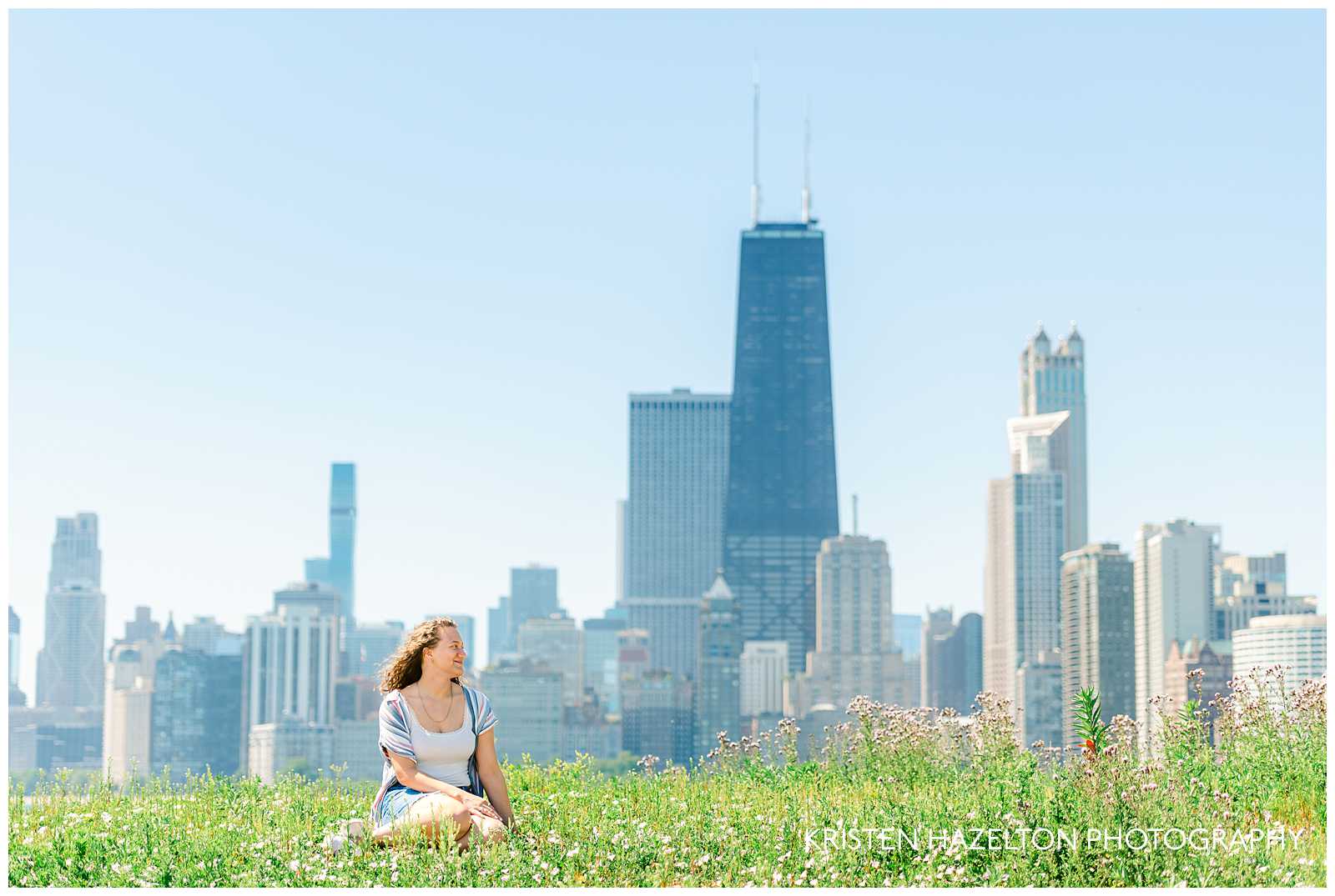 Girl with brown curly hair and white tank top sitting in wildflowers in front of the Chicago skyline