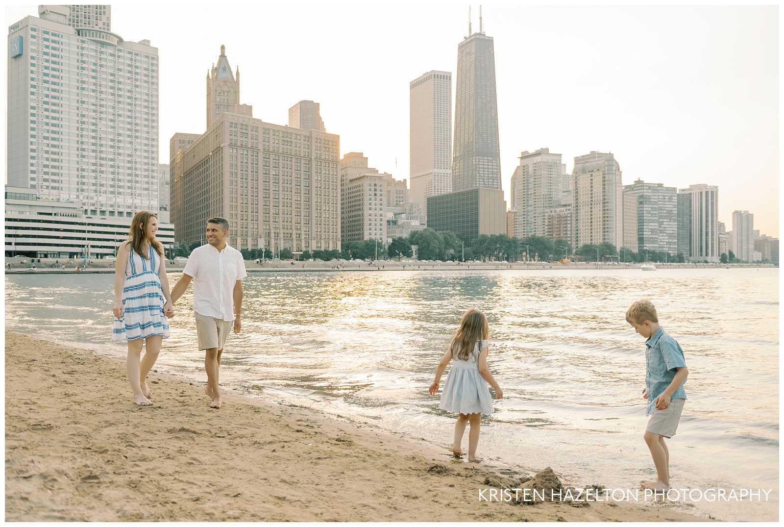 Father in white shirt and khaki shorts and mother in white and blue striped sundress walking along the beach while their two children make a sandcastle. The Chicago skyline appears in the background of their beach family photos.