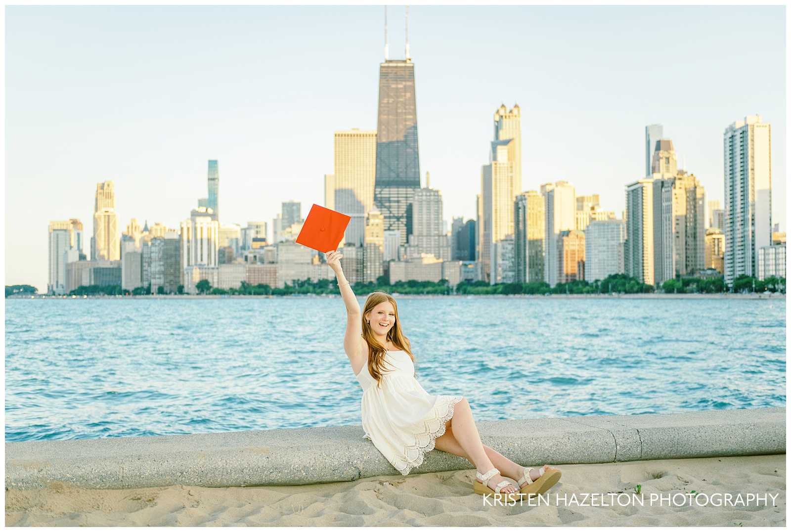 Girl with red hair wearing a white dress and holding her red graduation cap in the air. She is seated in front of the Chicago skyline at North Avenue Beach for her beach graduation photos