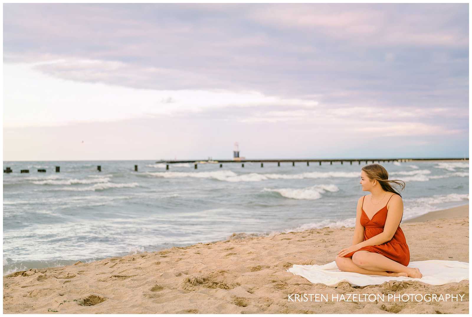 girl in a short red dress seated on a white blanket at the beach and looking out at a lighthouse at sunset. Beach senior pictures by Chicago senior photographer Kristen Hazelton