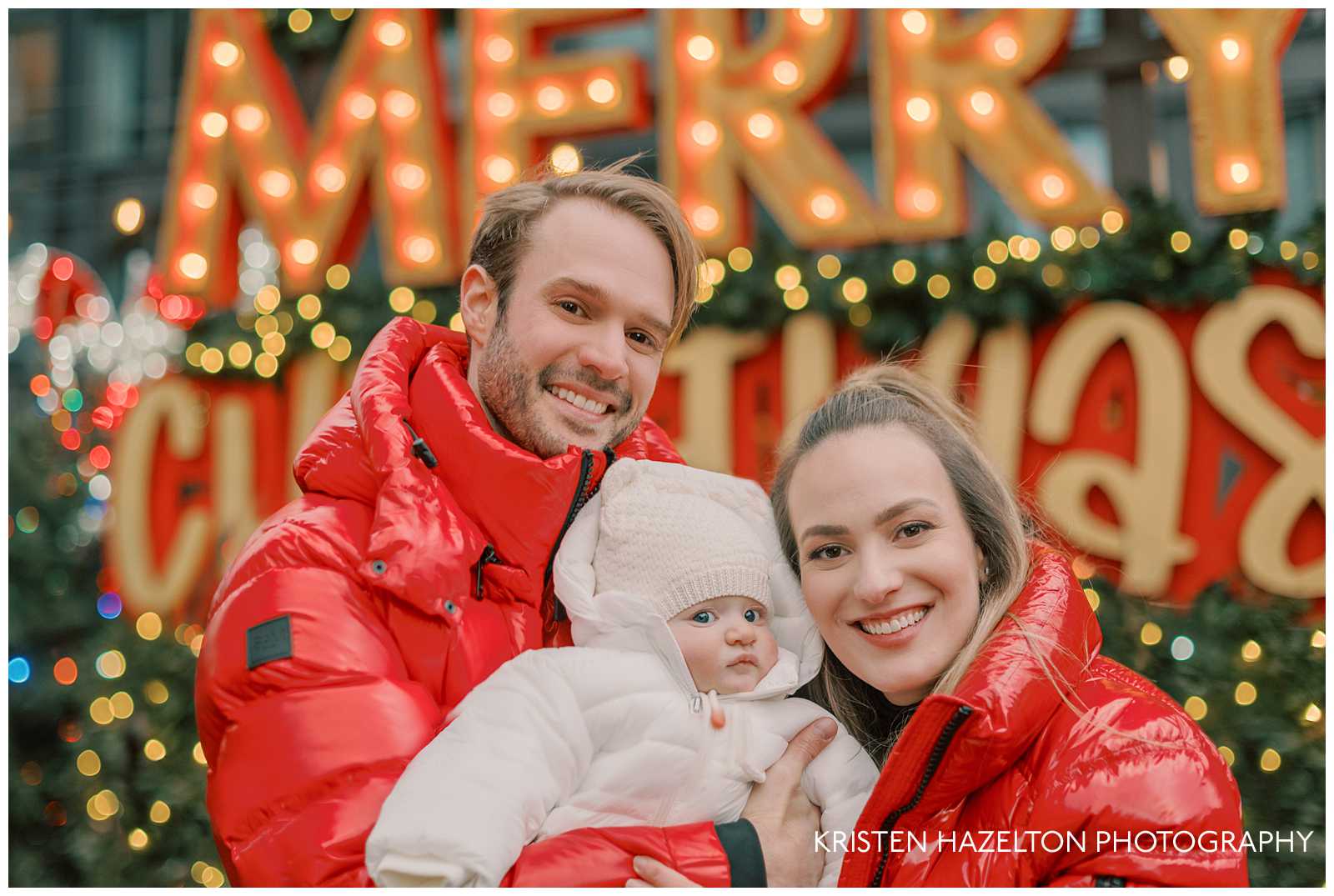 Family of three taking photos at the Jack Frost pop up in Fulton Market. The mom and dad are wearing red jackets while their baby is in a white snowsuit. They are standing in front of a sign that says Merry Christmas, with blurred holiday lights.