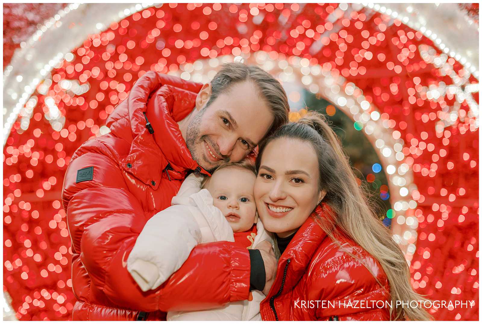 Vibrant family Christmas photos at the Jack Frost holiday pop up in Fulton Market. Mom and Dad wearing red jackets hugging their baby son who is wearing a white snowsuit in front of red and white christmas lights