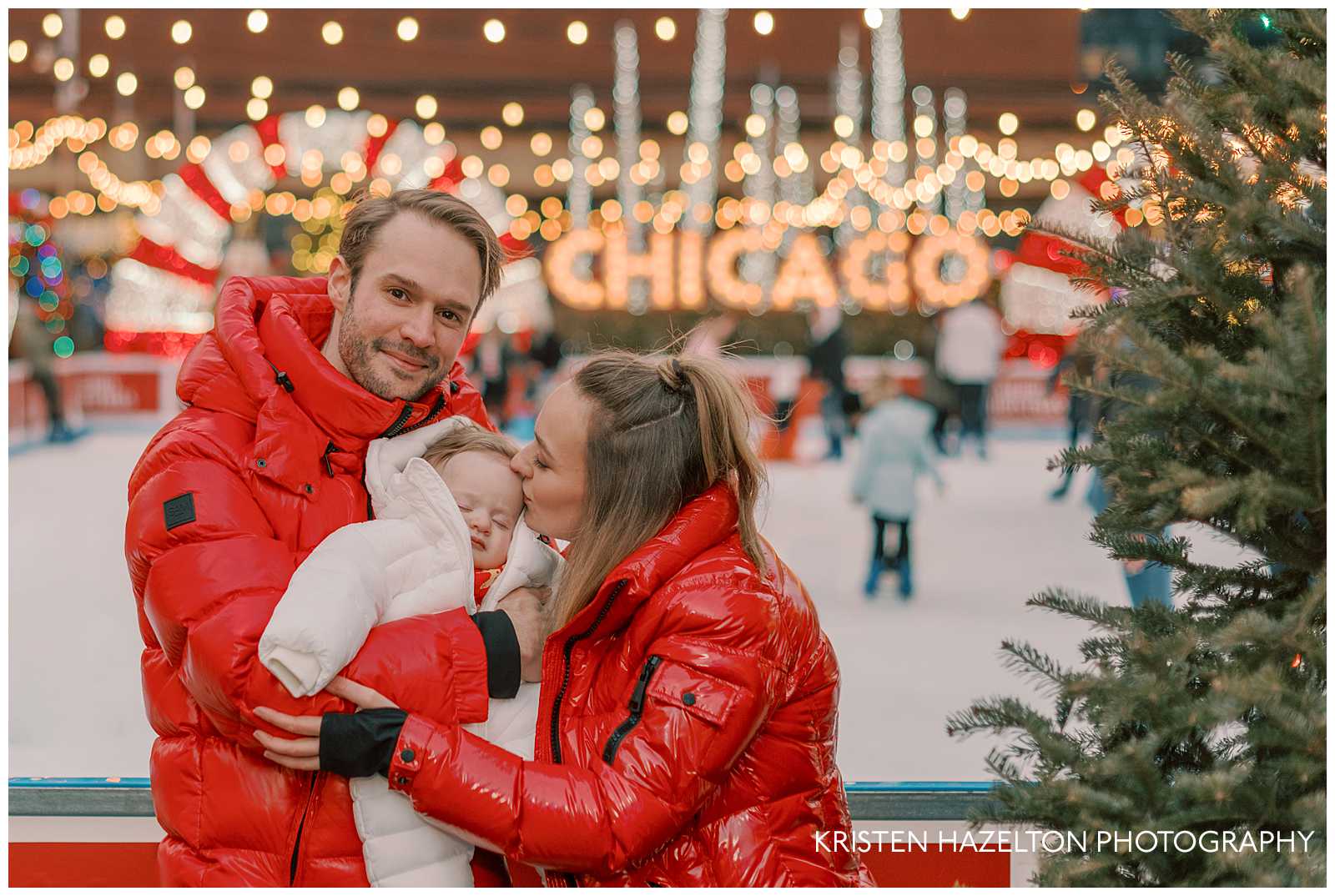 Young mom and dad wearing red jackets and holding their sleeping son for their holiday photos at the Jack Frost pop up in Chicago. They are standing in front of an ice skating rink with holiday lights and a Chicago light up sign in the background.