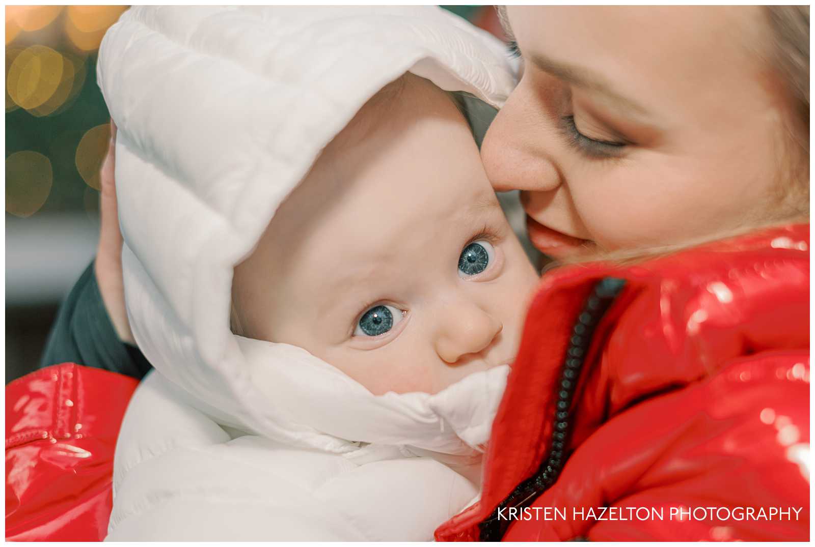 Closeup image of Mom in red jacket cuddling her baby son in a white snowsuit while he looks at Christmas lights for the first time. 