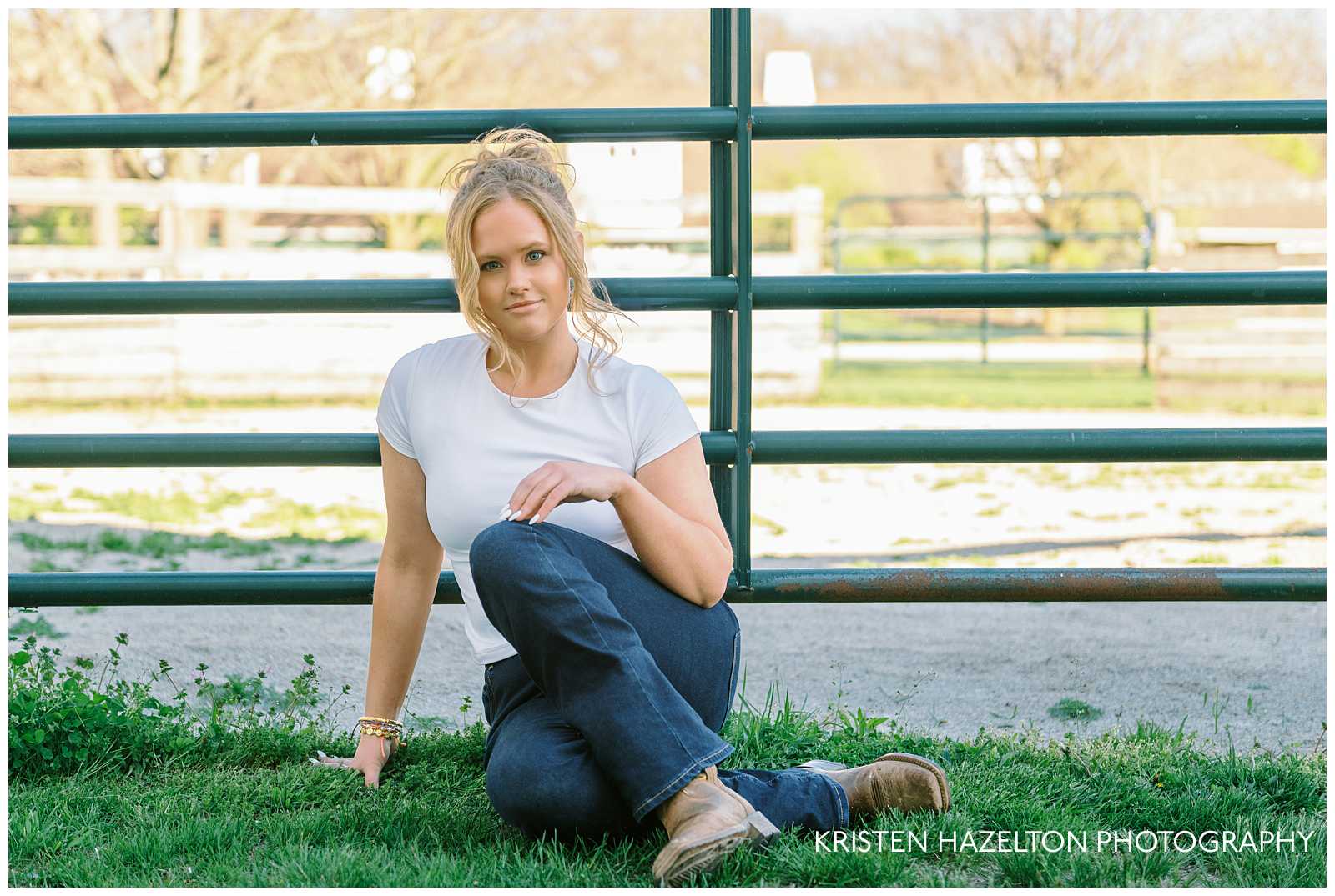 High school senior girl wearing a white t shirt and blue jeans seated in front of a farm gate at the Danada Equestrian Center in Wheaton IL