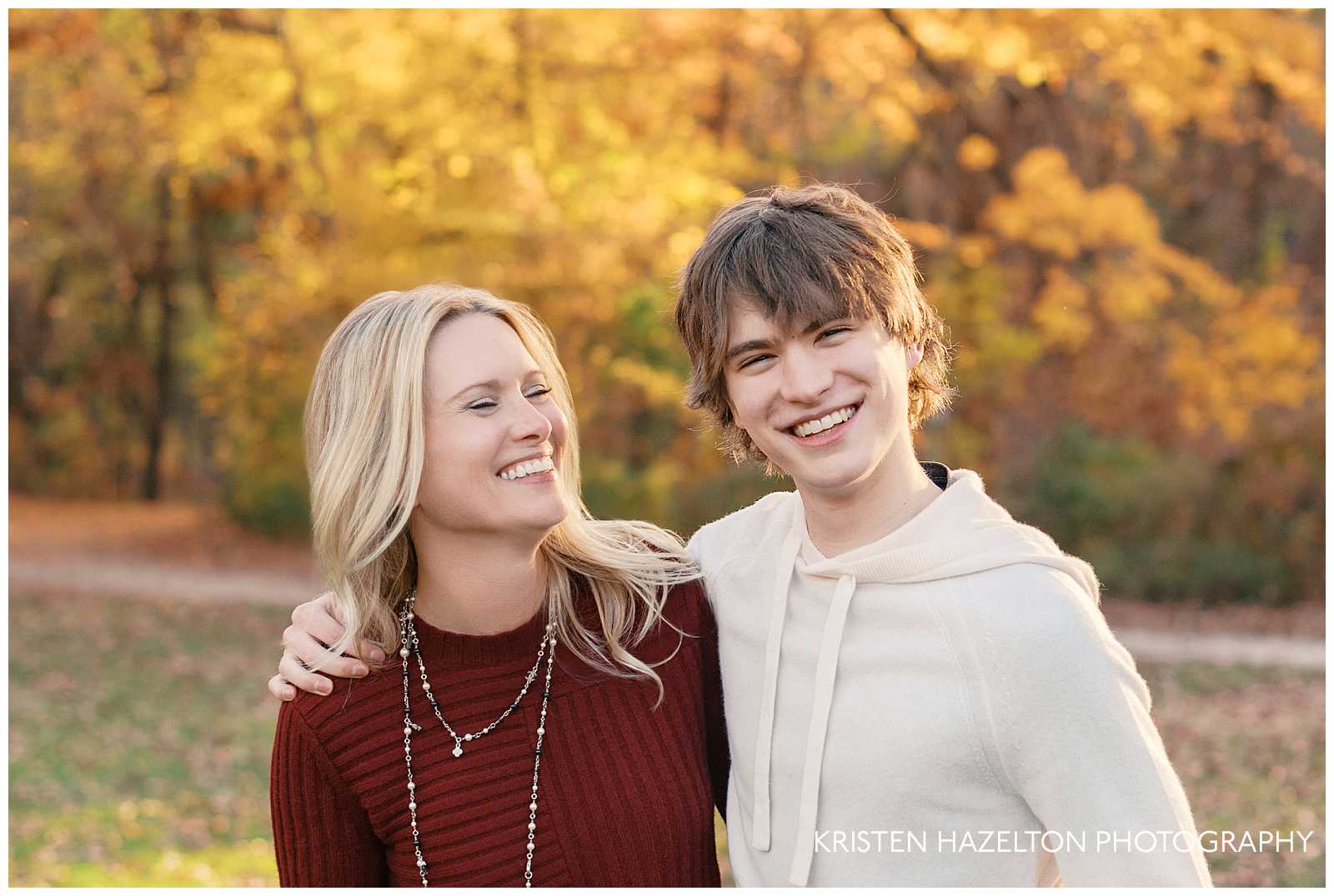 Mom in red sweater hugging her teenage son wearing a white sweatshirt. They are smiling in the woods with fall color in their photos by Evanston photographer Kristen Hazelton. 