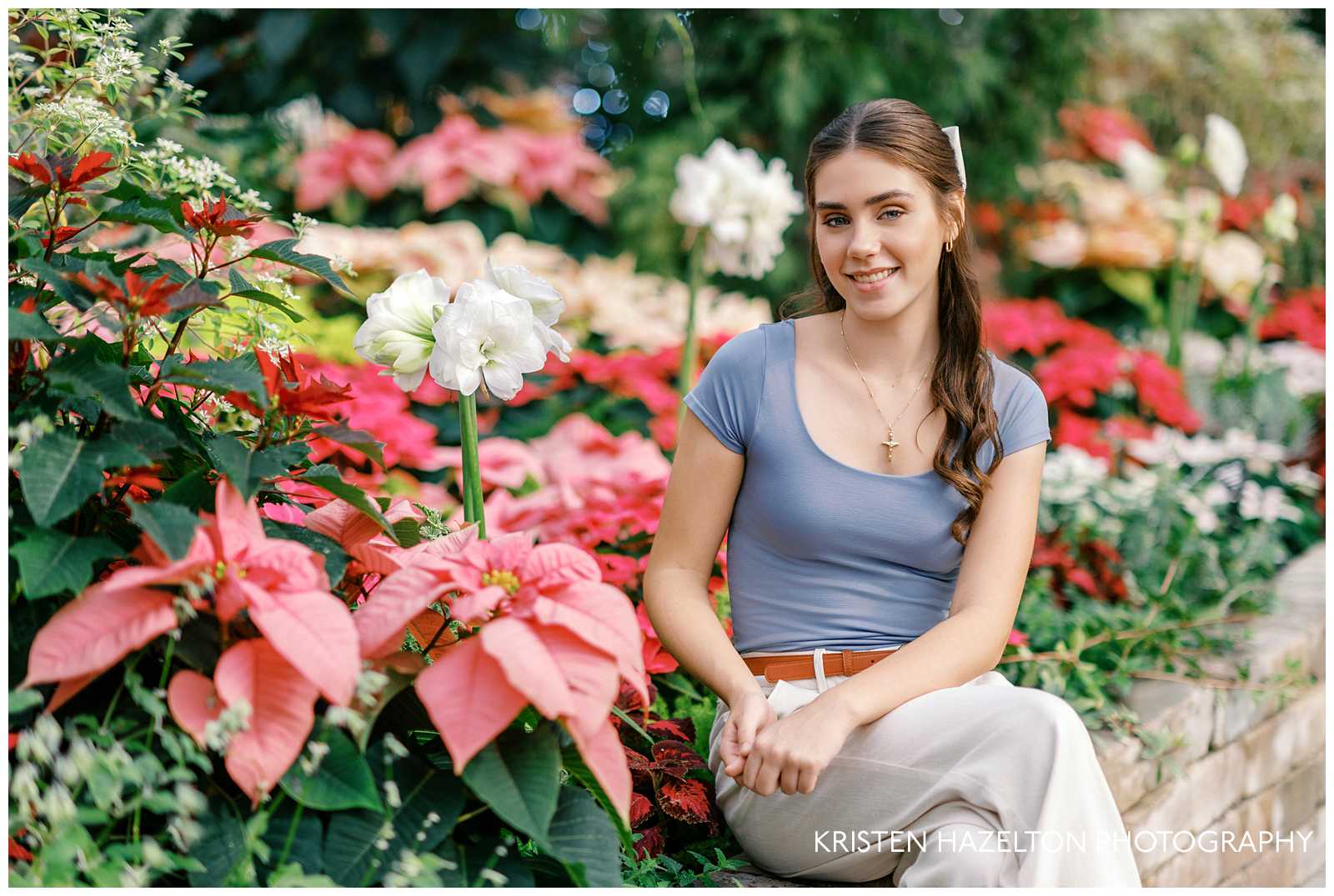 Christmas senior photos with poinsettias; a girl with a blue shirt and white pants sits next to a holiday greenhouse display 