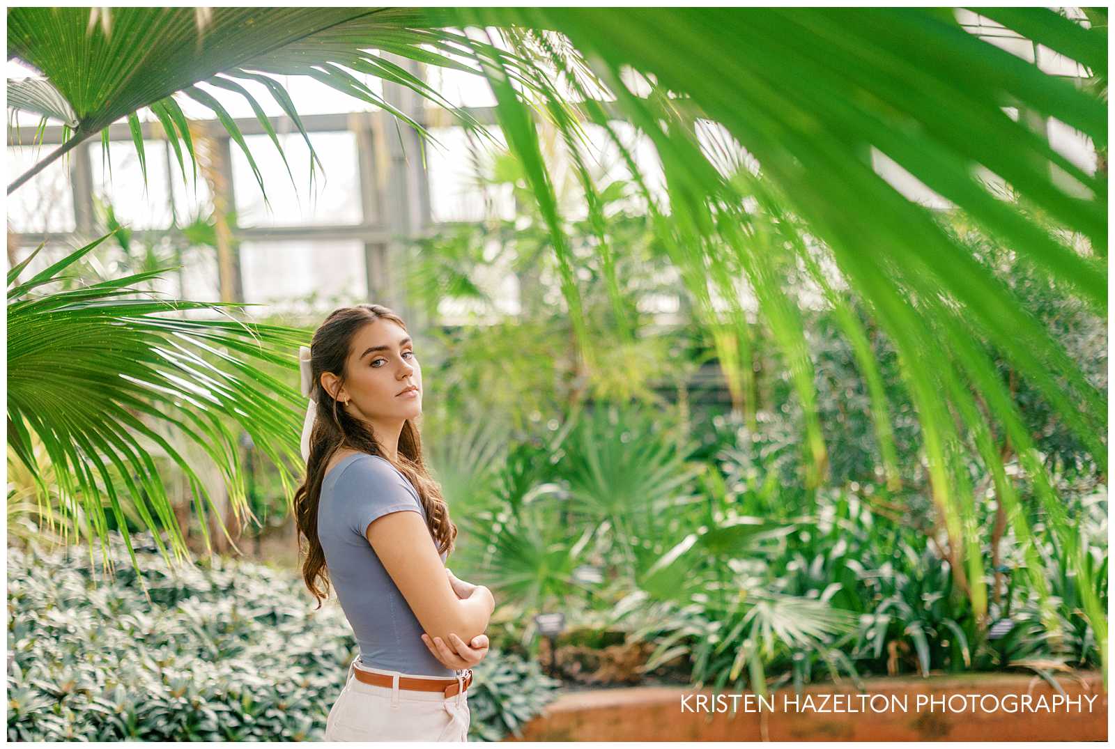 High school senior girl in blue top and white pants framed by a palm frond during her senior photos at the Garfield Park Conservatory in Chicago