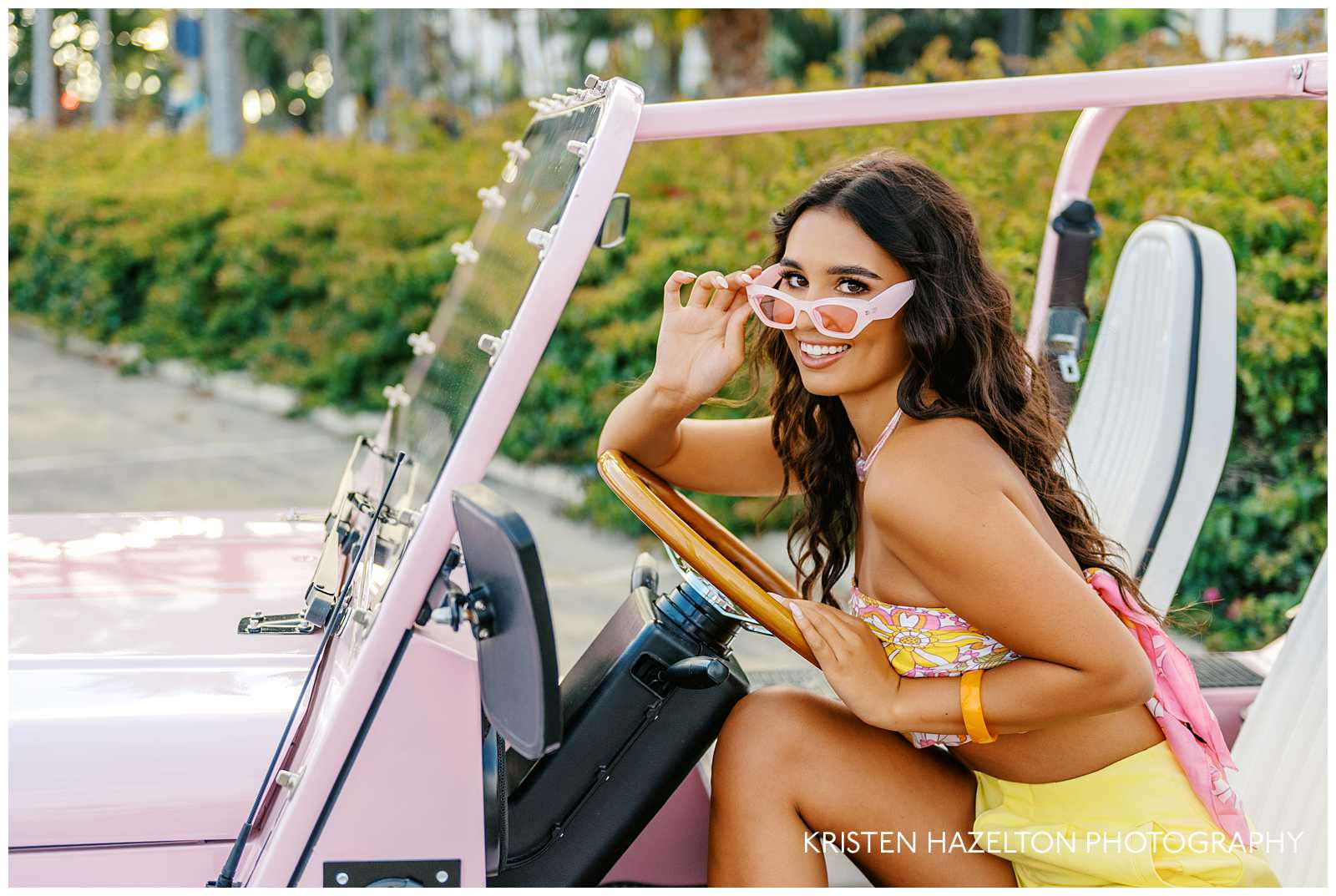 Girl with brown hair sitting in the driver's seat of a pink car and playing with her sunglasses for her Barbie photoshoot
