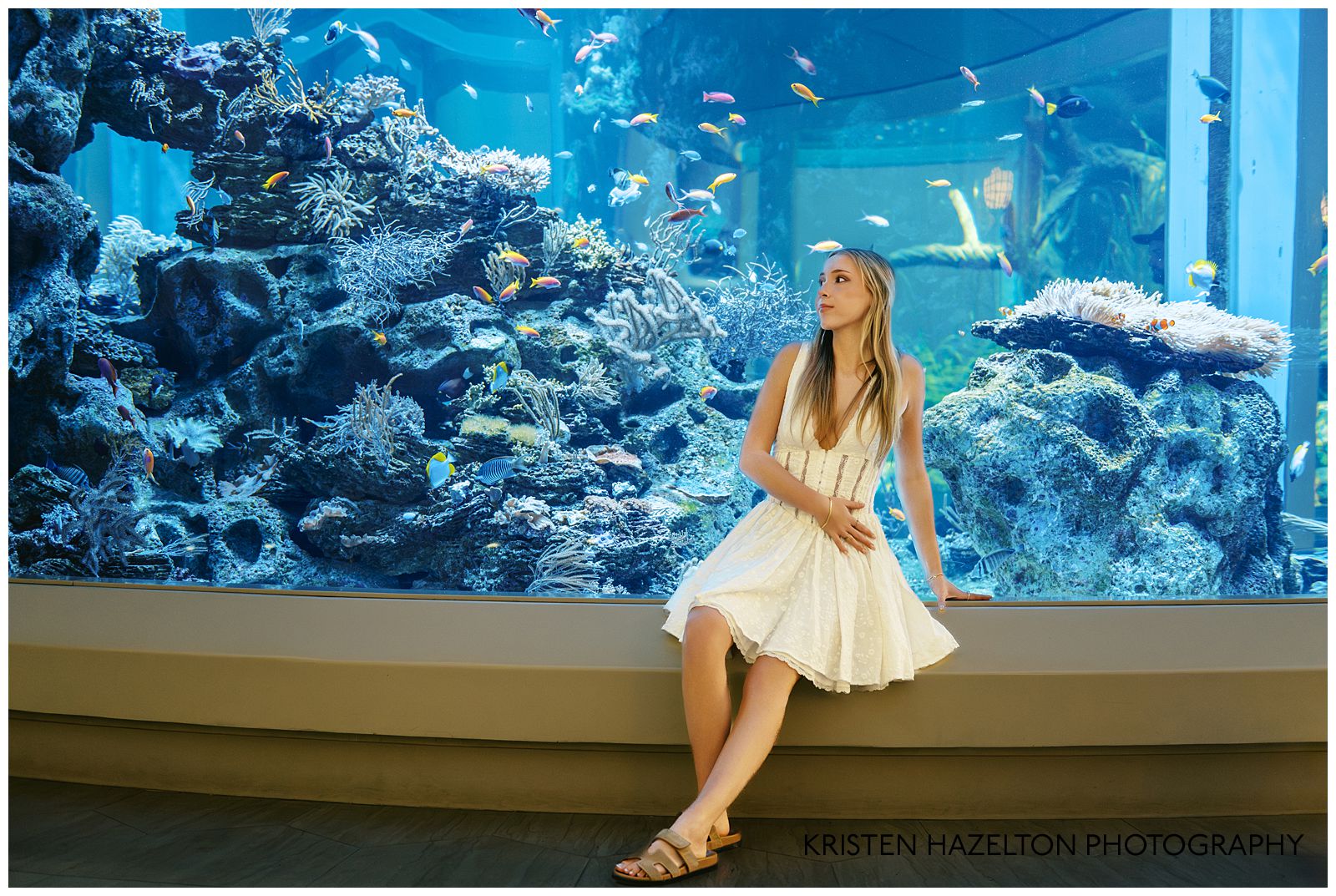 Girl in white dress seated next to a large tank of tropical fish for her aquarium photoshoot.