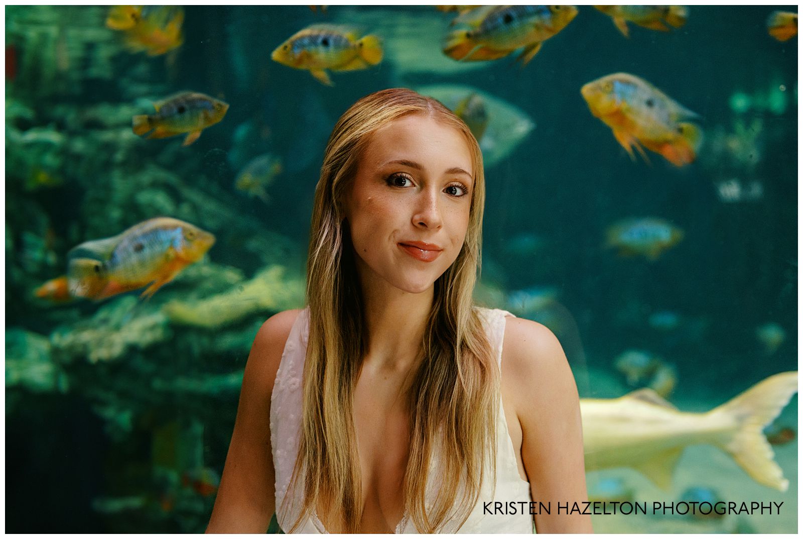 Girl in white dress seated next to a large tank of tropical fish for her aquarium photoshoot.