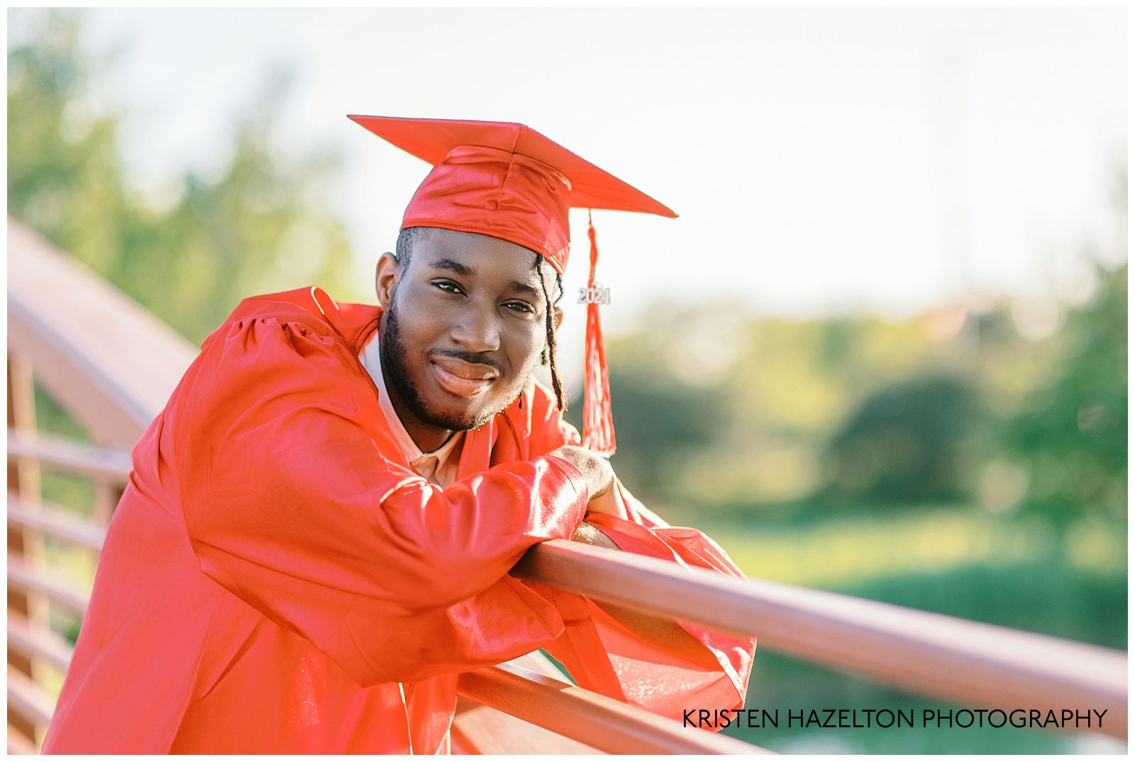 Male high school senior wearing a red cap and gown and leaning over the rail of a bridge for his portraits with Indiana senior photographer Kristen Hazelton